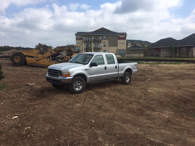 Silver Ford pickup truck parked on a dirt lot with construction equipment and buildings in the background.