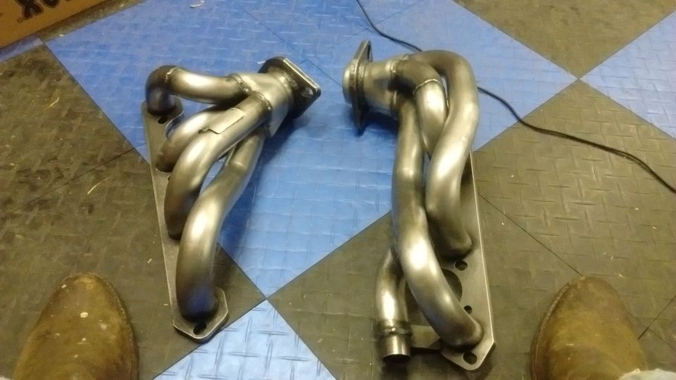 Two bare steel shorty exhaust headers standing on a blue and black diamond-plate floor