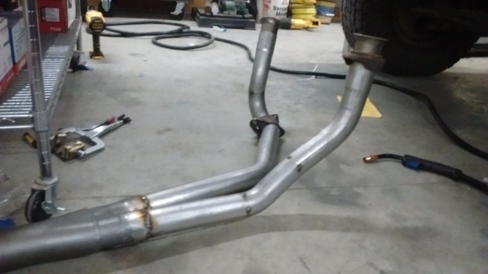 Custom exhaust system being welded on garage floor, with welding torch and vehicle tire visible.