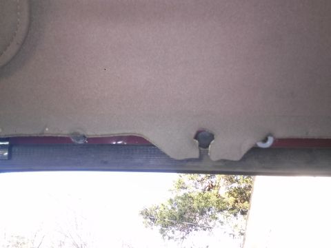 1996 Ford Bronco - The headliner of a vehicle is detached near the top of the windshield, revealing a hole and a white plastic clip. The maroon metal trim of…