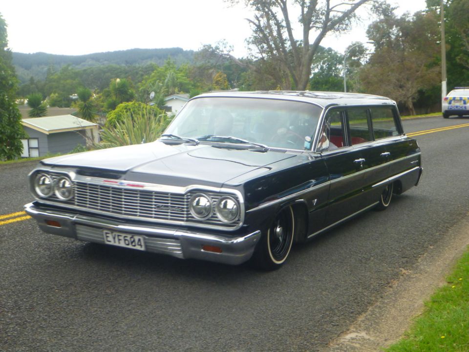 Black 1960s station wagon with whitewall tires driving on a road, police car in background.