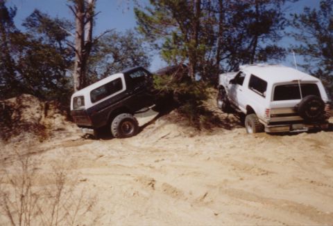 1995 Ford Bronco - Two lifted Ford Broncos on a sand hill in a pine forest: a blue-and-white bullnose-era Bronco climbing at a steep angle on the left, and a…