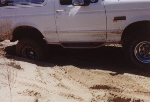 1995 Ford Bronco - Close side view of a white Ford Bronco buried to the axles in loose sand, front tire dug into a rut with churned tracks in the foreground.…