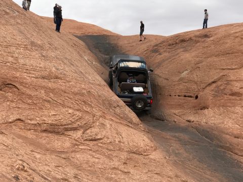 1991 Ford Bronco - A dark blue SUV is navigating a steep, rocky incline with several people standing on the surrounding red rock formations. The vehicle appea…