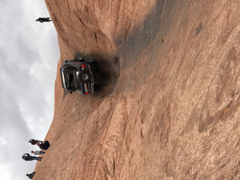 1991 Ford Bronco - A dark blue Jeep Wrangler is precariously balanced on its side on a steep, rocky incline, with several people watching from below.