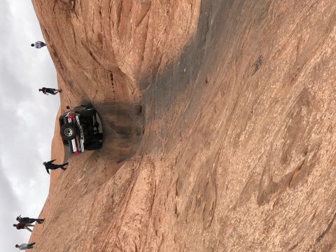 1991 Ford Bronco - A black Ford Bronco is precariously parked on a narrow ledge of a large rock formation, with several people standing around it on the cliff.