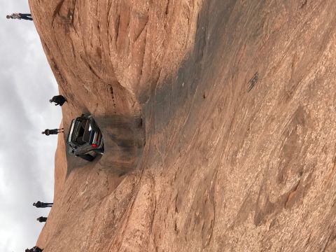 1991 Ford Bronco - A dark-colored SUV is precariously wedged into a narrow rock crevice on a steep sandstone cliff. Several people are standing on the cliff e…