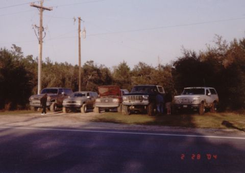 1995 Ford Bronco - A row of five lifted Ford trucks and Broncos parked nose-out on a grass shoulder beside a paved road, with people standing among them. The…