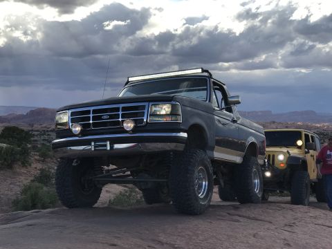 1991 Ford Bronco - A lifted dark-colored Ford pickup truck with a light bar on the roof is parked on a rocky terrain next to a yellow Jeep. The sky above is f…