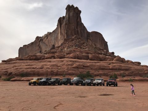 1991 Ford Bronco - A line of off-road vehicles, including several Jeeps and a yellow SUV, are parked on a dirt surface in front of a large, layered red rock f…