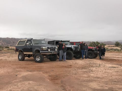 1991 Ford Bronco - A group of people pose with a lifted dark blue Ford Bronco and two lifted Jeep Wranglers in a desert landscape under a cloudy sky.