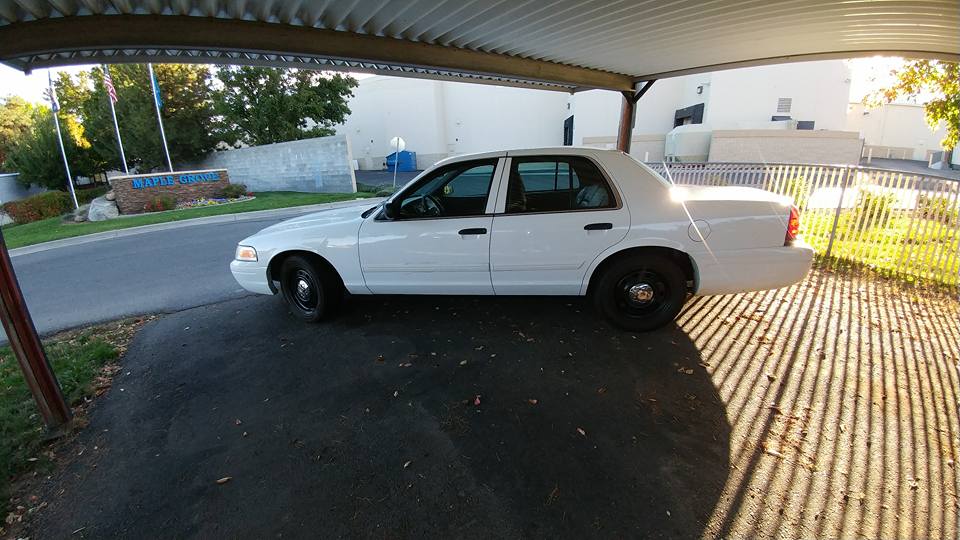 White sedan, likely a Ford Crown Victoria, parked under a carport. Shadows from the carport roof fall across the ground
