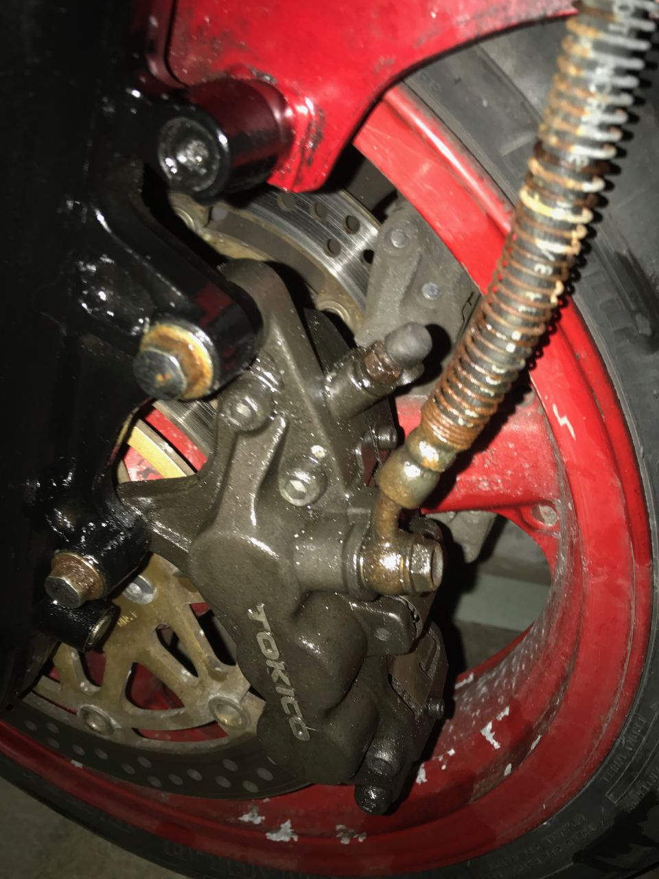Tokico front brake caliper and drilled rotor on a red motorcycle wheel with rusty spring-wrapped brake line