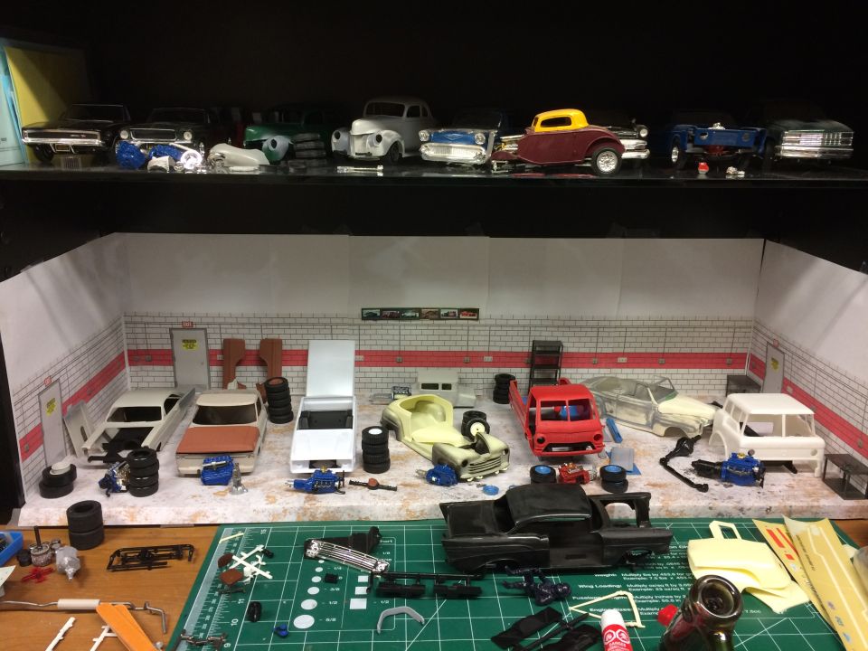 Model car garage diorama with several unfinished car models and parts on a workbench