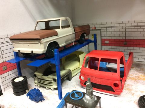 1989 Ford Bronco - Model car garage scene with unfinished trucks on a lift and various parts on the floor.
