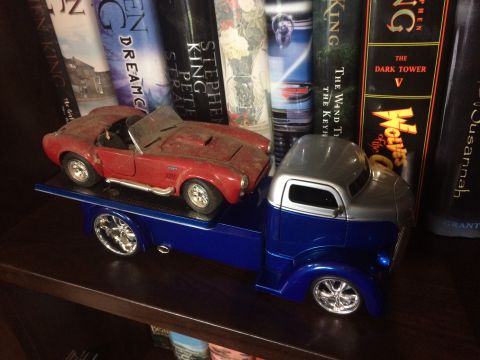 1989 Ford Bronco - A model of a red convertible sports car is loaded onto a blue tow truck, displayed on a shelf with books.