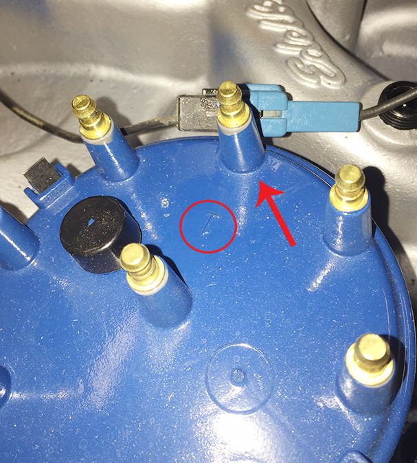 Close-up of a blue distributor cap with brass terminals. A red arrow points to a small 'T' marking