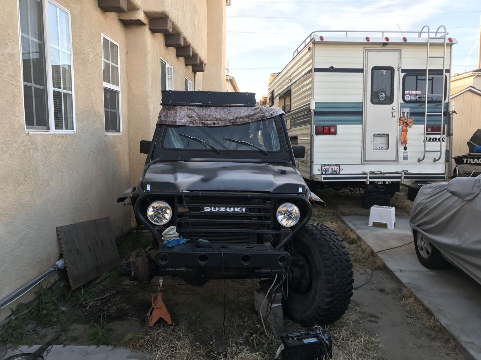 Front view of a black 1987 Suzuki Samurai with a roof rack, parked next to an RV