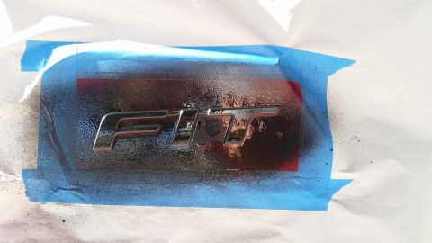2015 Honda Fit - A chrome FIT emblem is taped off and being spray painted, with overspray visible on the blue masking tape.