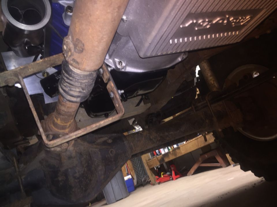 Underside view of a vehicle's drivetrain, showing the driveshaft, differential, and part of the transmission. There's a logo on a metal plate