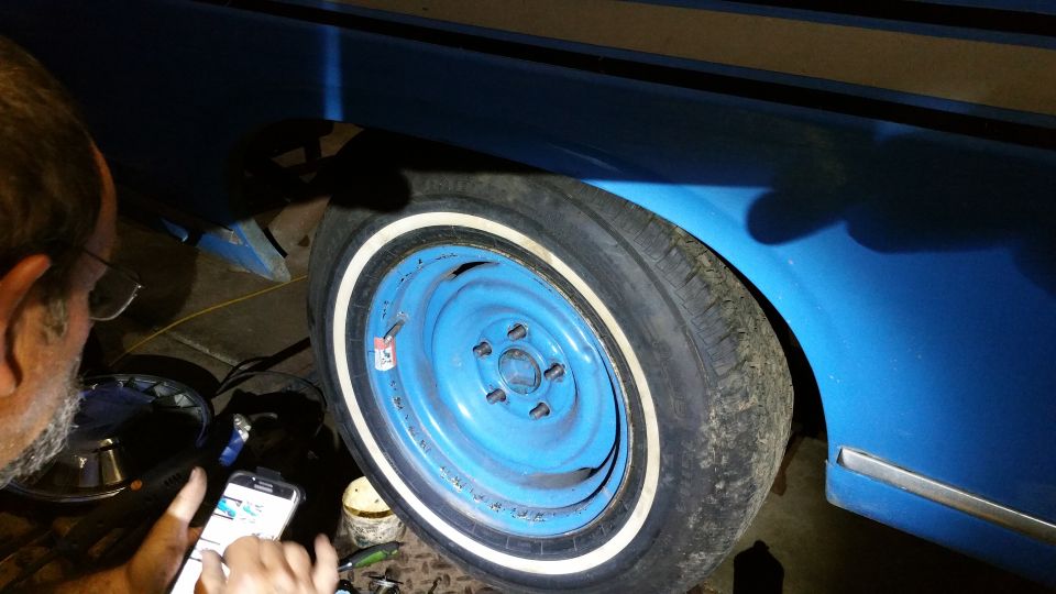 Man looking at phone next to a blue car's wheel and fender. The wheel is painted blue with a white stripe tire