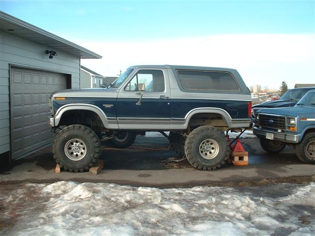 Lifted blue and silver 1980s Ford Bronco on blocks in a snowy driveway