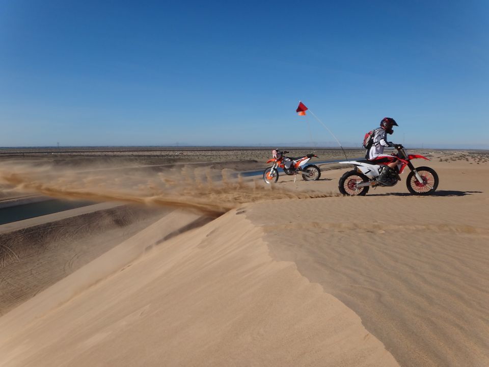 Two dirt bikes riding on sand dunes, kicking up dust. One rider has a red flag