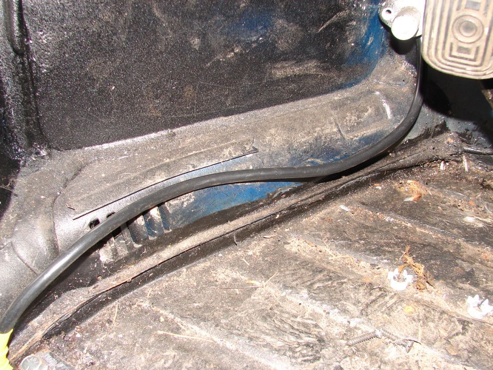 Interior floor pan of a vehicle, showing pedals and wiring. The area is dusty and shows some rust