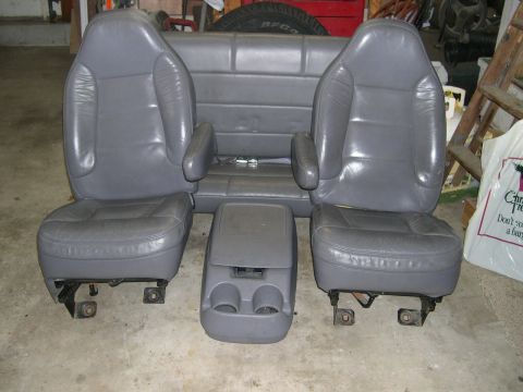 1994 Ford Bronco - Set of gray leather vehicle seats removed and staged on a garage floor: two bucket seats with fold-down armrests on their brackets, a match…
