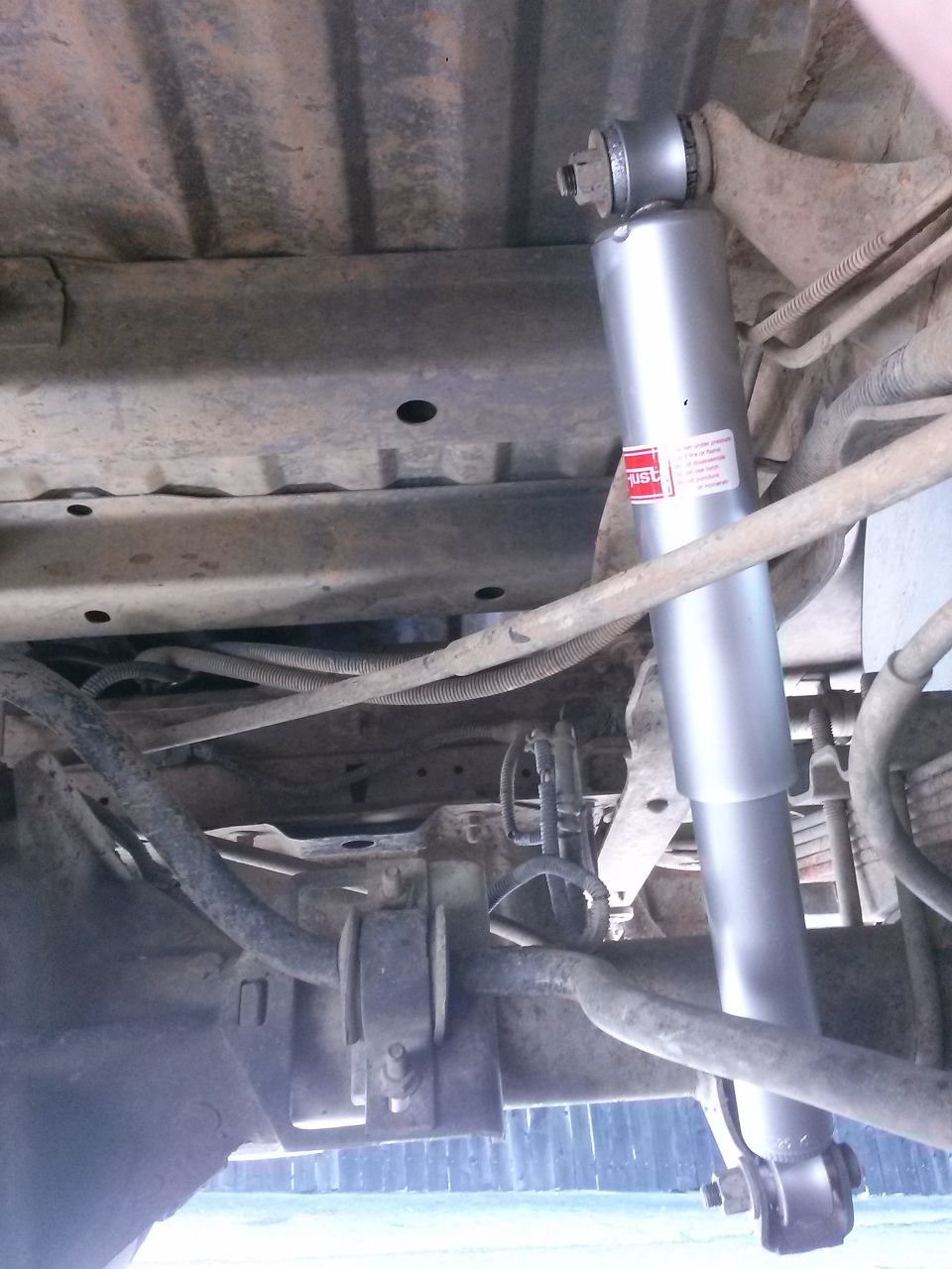 Underside of a vehicle showing a silver shock absorber with a red and white label, attached to the rear axle and frame