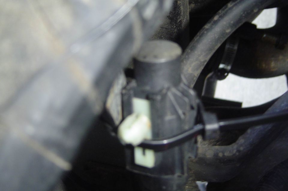 Close-up of a small black sensor with white connector mounted in a dusty engine bay