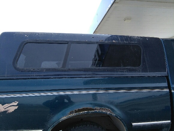 Side view of a dark green pickup truck with a dark blue topper. Some rust is visible on the rear fender