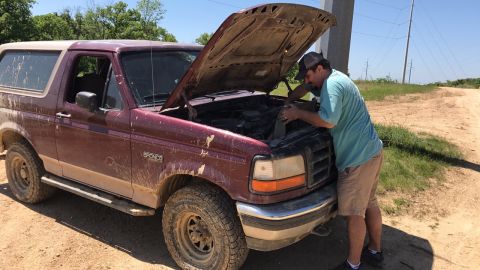 1996 Ford Bronco - Man works on the engine of a dirty 1996 Ford Bronco on a dirt road.