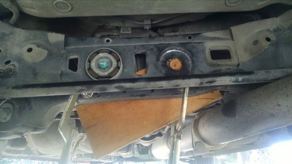 Undercarriage of a vehicle with a wooden block and clamps supporting a metal component. Exhaust pipes are visible