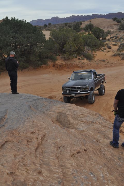 1991 Ford Bronco - A dark gray Ford F-Series pickup truck navigates a rocky, off-road trail with two people observing.