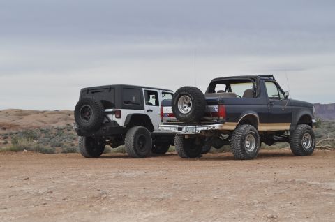 1991 Ford Bronco - Two modified SUVs, a 1991 Ford Bronco and a Jeep Wrangler, are parked side-by-side on a dirt track.