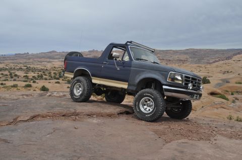 1991 Ford Bronco - A lifted dark blue Ford Bronco with tan side stripe and off-road tires sits on a rocky desert terrain under a cloudy sky.