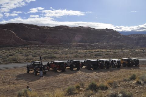 1991 Ford Bronco - Several off-road vehicles are parked in a line on a dirt lot with a mountainous desert landscape in the background.