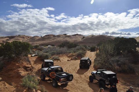 1991 Ford Bronco - Several off-road vehicles navigate a dusty trail through a desert canyon under a cloudy sky.