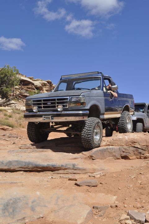1991 Ford Bronco - This lifted Ford Bronco navigates a challenging rocky trail under a bright blue sky.