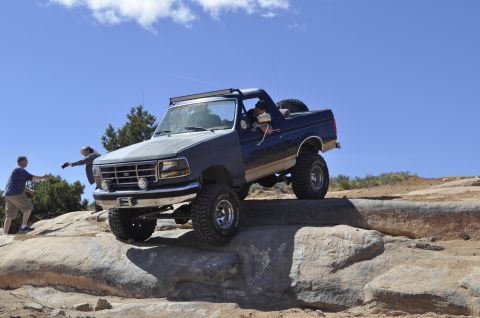 1991 Ford Bronco - A lifted Ford Bronco navigates a rocky obstacle on an off-road trail under a clear blue sky.