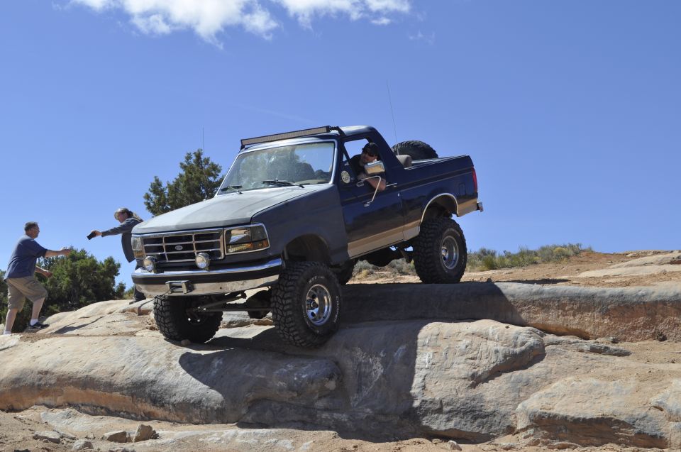 Ford Bronco climbing rocks on a trail, driver visible, two people watching