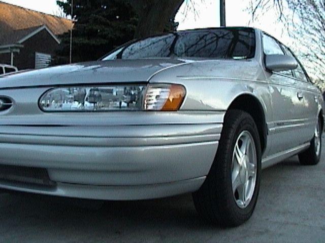 Front corner of a silver Ford Taurus sedan parked in a driveway, close on the headlight and wheel