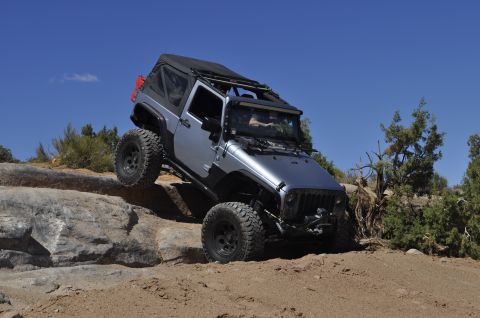 1991 Ford Bronco - A silver Jeep Wrangler navigates a rocky off-road trail under a clear blue sky.