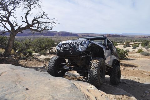 1991 Ford Bronco - A heavily modified Jeep Wrangler navigates a challenging rocky off-road course under a partly cloudy sky.