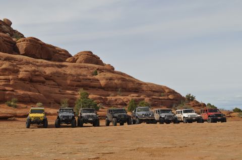 1991 Ford Bronco - A group of Jeeps are lined up in a desert environment with large red rock formations under a cloudy sky.