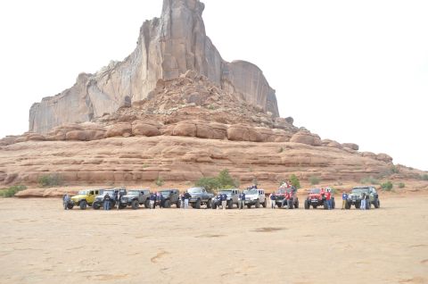 1991 Ford Bronco - A convoy of off-road vehicles is parked in a desert landscape, with a towering rock formation dominating the background.