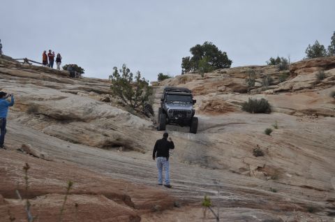 1991 Ford Bronco - A Jeep Wrangler navigates a steep, rocky incline in a desert landscape, observed by onlookers.