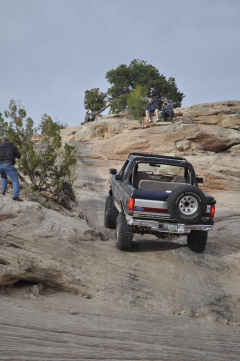 1991 Ford Bronco - A dark blue Ford Bronco navigates a steep, rocky incline with a spare tire mounted on the back.
