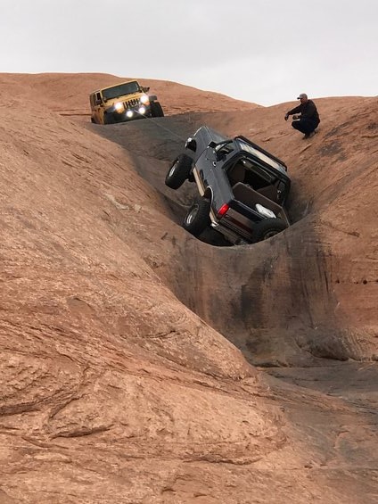 1991 Ford Bronco - A dark blue SUV is being towed up a steep, rocky incline by a yellow Jeep, with a person watching from the ridge.